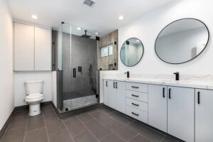 Luxury bathroom with round mirrors above a wooden vanity, glass shower enclosure, premium toilet, ambient lighting, and view of outdoor space.